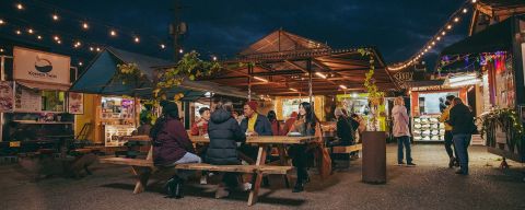 people seated at picnic tables talk and eat with food carts in the background