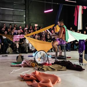 a person sits on a stool in front of an audience with colorful clothing strewn about the stage