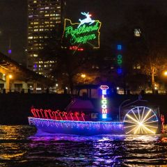 a boat festively decorated with holiday lights cruises along a river beneath the Portland White Stag sign