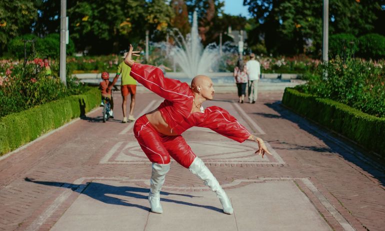 a person in bright clothes and high-heeled boots vogues in front of a park fountain, surrounded by manicured rose bushes