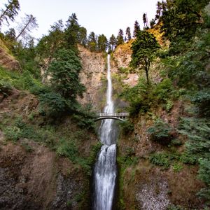 sunset hits the top of the mountain where a large waterfall bursts through the trees and cascades down the hillside, with a small bridge overlook
