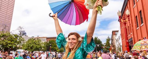 a smiling person at a parade holds a rainbow umbrella and a bouquet of flowers above their head