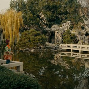 A woman holds a camera to her eyes in a traditional Chinese garden with a pond, pavilion and trees.