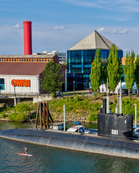 an exterior view of the Oregon Museum of Science and Industry (OMSI) beside the Willamette River with a huge black submarine docked by a pier