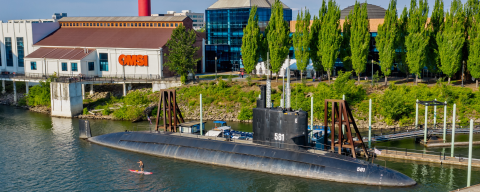 an exterior view of the Oregon Museum of Science and Industry (OMSI) beside the Willamette River with a huge black submarine docked by a pier